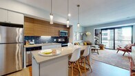 This contemporary single-floor residence offers a thoughtfully designed open-concept layout that seamlessly connects the kitchen, dining, and living areas. The kitchen is a standout feature, boasting two-tone cabinetry with warm wood upper cabinets and crisp white lower units, a bold navy blue subway tile backsplash, stainless steel appliances, and a central island with quartz countertops and pendant lighting overhead. Light wood flooring runs throughout, adding warmth and continuity to the space. The living room is bright and inviting, anchored by large floor-to-ceiling sliding glass doors that open to a private balcony, offering expansive urban views and generous natural light. A curated gallery wall and stylish arc floor lamp contribute to the room's modern-bohemian character. The bedroom continues the airy aesthetic, featuring large windows with city views, soft neutral tones, and a wall-mounted display shelf. The bathroom is finished with clean, modern lines, including a white floating vanity with ample storage, a quartz countertop, and contemporary hardware. Throughout the residence, the design balances modern minimalism with warm, eclectic accents, creating a cohesive and comfortable living environment.