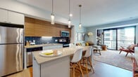 This contemporary single-floor residence offers a thoughtfully designed open-concept layout that seamlessly connects the kitchen, dining, and living areas. The kitchen is a standout feature, boasting two-tone cabinetry with warm wood upper cabinets and crisp white lower units, a bold navy blue subway tile backsplash, stainless steel appliances, and a central island with quartz countertops and pendant lighting overhead. Light wood flooring runs throughout, adding warmth and continuity to the space. The living room is bright and inviting, anchored by large floor-to-ceiling sliding glass doors that open to a private balcony, offering expansive urban views and generous natural light. A curated gallery wall and stylish arc floor lamp contribute to the room's modern-bohemian character. The bedroom continues the airy aesthetic, featuring large windows with city views, soft neutral tones, and a wall-mounted display shelf. The bathroom is finished with clean, modern lines, including a white floating vanity with ample storage, a quartz countertop, and contemporary hardware. Throughout the residence, the design balances modern minimalism with warm, eclectic accents, creating a cohesive and comfortable living environment.