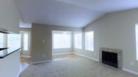 This single-story space showcases a thoughtfully designed open living area that balances comfort and functionality. The main living room features plush wall-to-wall carpeting and is anchored by a tile-surround fireplace with a sleek black insert, serving as a natural focal point within the room. Neutral, warm-toned walls throughout create a welcoming and versatile backdrop suitable for a variety of décor styles.

A distinctive step-down area adjacent to the living room transitions to hardwood flooring, adding visual interest and spatial definition. This sunlit nook is framed by multiple large windows fitted with white horizontal blinds, allowing abundant natural light to fill the space while maintaining privacy. A ceiling fixture in this area provides additional illumination.

White baseboards and trim throughout the space offer clean contrast against the neutral walls, contributing to a polished overall appearance. The open layout between the living and adjoining areas promotes a sense of spaciousness and ease of movement. The combination of the fireplace, generous natural light, and varied flooring materials gives this space a practical yet inviting character well-suited for everyday living.