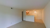 This single-floor space is designed with an open-concept layout that connects the living area to the kitchen. The living room is finished with wall-to-wall beige carpeting, which adds warmth and continuity throughout the space. Neutral white walls and a standard drop ceiling contribute to a clean, simple aesthetic, while a ceiling-mounted ventilation duct ensures climate control comfort.

A half-wall partition separates the living area from the kitchen, forming a pass-through counter that enhances the open feel while defining each functional zone. The kitchen features warm wood-toned upper cabinetry and includes a refrigerator, giving it a traditional, practical character. A ceiling fan is positioned in the adjoining area near the kitchen, promoting air circulation throughout the connected spaces.

A hallway visible along one wall leads to additional rooms, while a doorway on the opposite side of the kitchen area provides further access within the unit. The overall layout is straightforward and functional, making efficient use of the available space while maintaining an open, airy feel throughout the main living and kitchen areas.