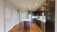 This single-floor residence showcases a cohesive, modern design throughout. The kitchen serves as a central highlight, featuring rich dark-espresso cabinetry that extends to the ceiling, a classic white subway tile backsplash, and a full suite of stainless steel appliances including a gas range, microwave, refrigerator, and dishwasher. A central island with a clean white countertop offers additional prep space and is illuminated by stylish pendant lighting overhead, while recessed lighting ensures the area remains bright and functional. Warm-toned hardwood-style flooring flows seamlessly from the kitchen into the open living area, creating a sense of visual continuity and spaciousness. The living space is generously proportioned, finished with neutral gray walls and large windows that allow natural light to filter in. The bedroom is carpeted for comfort and includes a ceiling fan and a wide window offering natural light. The bathroom is cleanly appointed with large-format light tile flooring, a dark vanity with a white countertop, a framed mirror, and bar lighting. Throughout the unit, neutral tones and consistent finishes establish a polished, contemporary ambiance.