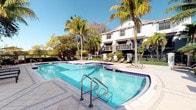 This inviting property showcases a beautifully appointed outdoor amenity area as its centerpiece. The sparkling swimming pool, trimmed with elegant blue tile accents and equipped with chrome entry rails, is the focal point of the outdoor space. A generously sized pool deck, finished in light-toned pavers, offers ample room for lounging and entertaining, lined with cushioned chaise lounge chairs and shaded umbrella seating for comfort under the Florida sun. Towering palm trees and lush tropical landscaping frame the area, creating a serene, resort-like atmosphere. The backdrop features a multi-story building with a classic Mediterranean-influenced architectural style, characterized by a crisp white stucco exterior, arched window detailing, and private balconies with wrought iron railings. A secondary spa or splash area is visible adjacent to the main pool, adding to the leisure offerings. The surrounding greenery and mature trees provide natural shade and privacy, enhancing the tranquil outdoor environment. The property's single-floor layout is complemented by these thoughtfully designed communal outdoor spaces, making it well-suited for those who value relaxed, resort-style living.