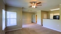This single-floor residence offers a thoughtfully designed layout with a welcoming and functional aesthetic. The main living area is generously sized, featuring plush wall-to-wall carpeting that adds warmth underfoot. Crown molding lines the ceilings, lending a refined architectural touch, while a ceiling fan with integrated lighting provides both comfort and illumination. A two-tone wall treatment, with a chair rail dividing the upper and lower sections, adds classic detailing to the interior.

Natural light filters in through a window fitted with horizontal blinds, brightening the space. Recessed track lighting provides additional overhead illumination, ideal for accent or task lighting. A half-wall partition with a granite or stone countertop edge hints at a connected kitchen or breakfast bar area, offering a sense of openness between living and culinary spaces.

Toward the rear of the main room, a doorway leads to what appears to be a private bedroom with an en-suite bathroom and walk-in closet visible beyond. Clean white trim and neutral green-toned walls create a calm, cohesive palette throughout, making this a move-in-ready space well-suited for everyday living.