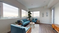 This single-floor unit showcases a thoughtfully designed interior with a contemporary aesthetic and mid-century modern influences throughout. The living area is a standout space, defined by an expansive bank of large windows along two walls that frame sweeping city and hillside views, flooding the room with natural light. Dark hardwood-style flooring runs throughout, providing a warm contrast to the crisp white walls and ceilings.

The kitchen is efficiently laid out in an L-shape configuration, featuring flat-front cabinetry in a muted slate-blue tone, a white tile backsplash with under-cabinet lighting, light stone countertops, and a full suite of stainless steel appliances including a French door refrigerator, gas range, built-in microwave, and dishwasher.

The bedroom is a calm, well-lit retreat with a large picture window offering scenic outdoor views and access to what appears to be an outdoor balcony area. Neutral walls and the consistent hardwood-style flooring maintain a cohesive feel throughout.

The bathroom is cleanly finished with neutral tile flooring, a combination tub and shower with glass sliding doors, a wall-mounted vanity with wood cabinetry, and a large mirror.