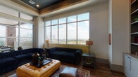 Located within the Hardy Yards development in Houston, TX, this stylish amenity lounge offers an elevated living experience with thoughtfully curated design details throughout. The space is anchored by an expansive wall of floor-to-ceiling grid-pane windows that frame sweeping, unobstructed views of the Houston skyline and surrounding landscape, flooding the interior with abundant natural light.

The main lounge area features rich hardwood flooring and a tray ceiling with warm wood accents and recessed lighting, creating a refined yet welcoming atmosphere. Plush navy blue tufted Chesterfield sofas and a cognac leather ottoman provide sophisticated seating arranged around a central coffee table, complementing the upscale aesthetic.

The reception and entry corridor showcases a striking combination of patterned tile and hardwood flooring, illuminated by elegant crystal chandelier pendants and set against deep teal accent walls. This transitional zone connects seamlessly to the seating area, with elevator access and a welcoming arrangement of accent chairs.

Large glass French doors open directly onto a generous outdoor terrace, where additional seating overlooks the cityscape, extending the living space outdoors. Bold contemporary artwork adorns the walls, adding a creative touch that reinforces the space's modern, urban character. Together, these elements create an inviting, upscale environment with a strong sense of place within the heart of Houston.