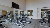 This fitness center is designed to provide a functional and comfortable workout environment. The space features soaring ceilings that lend an open, airy feel, while large windows allow natural light to fill the room and offer views of the surrounding exterior. A floor-to-ceiling mirror spans one wall, visually expanding the space and allowing users to monitor their form during exercise. The flooring consists of durable, patterned carpet suited for gym use. The cardio equipment lineup includes treadmills, elliptical trainers, and a stationary bike, all arranged to maximize usability and traffic flow. A large flat-screen television is mounted high on the wall, providing entertainment during workouts. Additional strength and resistance machines are visible along the perimeter, offering a well-rounded fitness experience. Informational signage is posted near the entrance, indicating facility rules and guidelines. The overall ambiance is clean, modern, and practical, reflecting a space designed with the active user in mind.