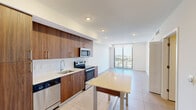 Located in the heart of Miami at 218 NW 8th Street, this contemporary unit offers a well-designed, single-floor layout that blends modern finishes with functional living. Light-toned porcelain tile flooring runs throughout, creating a clean and cohesive aesthetic across all spaces.

The kitchen is a standout feature, outfitted with rich walnut-finish cabinetry accented by gold hardware, a white subway tile backsplash, and a full suite of stainless steel appliances including a French door refrigerator, electric range, built-in microwave, and dishwasher. A freestanding dining table with a light countertop surface provides a casual dining option adjacent to the kitchen.

The open living area flows seamlessly from the kitchen toward a floor-to-ceiling glass sliding door that opens to a private balcony, offering sweeping views of the Miami skyline and surrounding cityscape. Natural light pours into the space, enhancing its bright, airy character.

The bedroom is generously proportioned, featuring a ceiling fan, large format tile flooring, and direct balcony access through its own glass slider. A closet with bi-fold doors provides convenient storage. The bathroom is elegantly appointed with marble-look wall tile surrounding the soaking tub, a floating walnut vanity with gold accents, and neutral tile flooring.