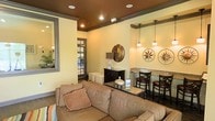 This single-floor space exudes a warm and welcoming atmosphere, thoughtfully designed with both comfort and style in mind. The main lounge area features a plush sectional sofa in earthy tones, accented with decorative throw pillows that invite relaxation. A rich, dark-painted ceiling adds depth and sophistication, complemented by recessed lighting that casts a warm glow throughout the room. Crown molding and carefully selected trim details reinforce the refined character of the space. A built-in counter area serves as a functional bar or workstation, outfitted with bar-height seating and illuminated by elegant pendant lights hanging from a rustic wooden beam. Decorative wall art, including a round metallic mirror and compass rose medallions, adds personality and visual interest to the warm yellow-toned walls. A dark wood console table with tasteful accessories anchors the transitional area between the lounge and the entryway. Large windows and a glass-paneled door allow natural light to filter in, offering glimpses of a bright outdoor area beyond. The hardwood flooring ties the entire space together, creating a cohesive and inviting environment suitable for both casual gatherings and quiet moments.