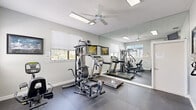 This fitness facility located at 1088 West Granada Boulevard in Ormond Beach, Florida, offers a practical and well-organized workout space designed to accommodate a range of exercise needs. The room features durable dark rubber flooring, which provides both traction and impact absorption during workouts. Neutral gray walls and white trim create a clean, bright atmosphere, complemented by natural light streaming in through multiple windows fitted with horizontal blinds.

The space is equipped with a multi-station strength training machine, a treadmill, and a stationary recumbent bike, offering options for both cardiovascular and resistance training. A large wall-mounted mirror spans one side of the room, visually expanding the space and allowing users to monitor their form. Motivational framed artwork is displayed on the walls, adding character to the environment. A ceiling fan with an integrated light fixture ensures air circulation and comfortable conditions during use.

Additional amenities include a wall-mounted television, a coat and key hook rack, and a hand sanitizer station, reflecting attention to hygiene and user convenience. The entry area provides a dedicated space to prepare before entering the workout floor. Overall, this gym delivers an efficient, well-maintained fitness environment suitable for a variety of exercise routines.