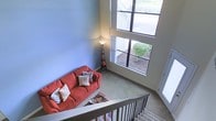 This well-appointed two-story residence offers an inviting and functional layout designed to maximize comfort and livability. The living area is a standout feature of the home, boasting dramatic double-height ceilings that lend a sense of grandeur and openness to the space. A large multi-pane window stretches upward toward the ceiling, flooding the room with natural light and offering pleasant views of the surrounding greenery outdoors. The soft, neutral-toned carpeting underfoot adds warmth and comfort to the living area, complementing the cool, light blue walls that create a calm and serene atmosphere. A staircase with a classic wood railing connects the two floors, serving as both a functional and architectural element that ties the home's design together. The upper level overlooks the living area below, reinforcing the home's open and connected feel. The overall ambiance of the space is warm and welcoming, with thoughtful design details — from the soaring ceilings to the generous window placement — working together to create a bright, comfortable, and visually appealing home environment.