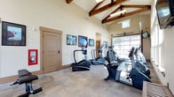 This single-floor amenity space is divided into two distinct areas: a fully equipped fitness center and a dedicated home theater room. The fitness center commands attention with its soaring vaulted ceiling adorned with exposed wooden beams, lending a warm, lodge-inspired aesthetic to the space. Large windows and an overhead garage-style door flood the room with natural light and offer views of the surrounding landscape. The facility features a comprehensive selection of True-brand cardio equipment, including ellipticals, recumbent bikes, and upright cycles, as well as a multi-station strength training machine. A wall-mounted flat-screen television, motivational artwork, and a wood-slatted bench complete the space. Carpeted flooring throughout provides comfort underfoot.

The adjacent screening room delivers a true cinematic experience, outfitted with rows of oversized, brown leather reclining chairs with built-in cup holders. A ceiling-mounted projector casts onto a large fixed projection screen flanked by built-in cabinetry and decorative floor plants. Recessed lighting and wall sconces create a warm, theater-like ambiance, while classic movie posters — including recognizable film franchise artwork — line the wainscoted walls, adding a nostalgic, curated character to the room.