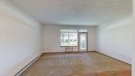 Located in Spokane, WA, this 2-bedroom, 2-bathroom single-floor residence offers a practical and well-organized layout. The open-concept living and kitchen area forms the heart of the home, where wood-look flooring transitions into carpeted living space. The kitchen features warm wood cabinetry, a tile backsplash, a curved breakfast bar countertop, and black appliances including a range, microwave, and refrigerator. The living room receives natural light through a large front-facing window alongside a entry door, with a baseboard heating system running along the wall. Both bedrooms are carpeted and neutrally finished, with the primary bedroom offering a generously sized walk-in closet equipped with wire shelving and hanging rods. The two bathrooms feature wood-look flooring, with one including a full tub-shower combination fitted with grab bars. A dedicated laundry room houses a full-size washer and dryer. The attached single-car garage provides secure parking and additional storage potential, with an automatic opener and drywall-finished interior. Textured ceilings and warm wood trim details are consistent throughout the home, lending a cohesive and comfortable feel to the overall space.