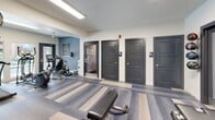 Located at 2800 S Syracuse Way in Denver, Colorado, this fitness center at the Arapahoe Club offers a comprehensive workout facility designed to support a wide range of exercise routines. The space is finished with a distinctive striped carpet in neutral tones of gray and beige, complementing the clean, modern aesthetic throughout.

The cardio area is positioned along a wall of large windows, flooding the space with natural light and providing views of the surrounding outdoor area, including a pool deck and mature trees. It is equipped with multiple treadmills, elliptical machines, and a recumbent bike, all from SportsArt and Matrix brands.

A dedicated strength training section features a full cable functional trainer system with a wide array of attachments, a dumbbell rack with a complete range of free weights, an adjustable workout bench, and a wall-mounted flat-screen television above a floor-to-ceiling mirror — ideal for monitoring form during exercise.

The facility also includes posted fitness center rules, indicating a well-managed and community-oriented environment. Toward the rear of the space, three gray paneled doors provide access to private restrooms — including an ADA-accessible option — and additional utility areas. Recessed and overhead panel lighting ensures consistent illumination throughout. The overall ambiance is clean, functional, and professional.