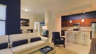 This single-floor space showcases a thoughtfully designed open-concept layout that connects the living area and kitchen, creating a cohesive and functional environment. The living area is anchored by a cream leather sofa accented with navy blue throw pillows, complemented by a matching navy armchair, lending the space a clean, nautical aesthetic. A glass-topped coffee table and a decorative tray featuring a starfish motif reinforce the coastal theme. A framed pier photograph adds a finishing artistic touch to the living space, while built-in shelving provides practical display and storage options.

The kitchen, distinguished by rich dark wood cabinetry set against a striking navy blue accent wall, transitions smoothly from the living area. Pendant lighting hangs above a bar-style counter with bar stools, ideal for casual dining. Stainless steel appliances and a tiled backsplash complete the kitchen's polished look. Light-toned tile flooring flows consistently throughout, unifying the space and enhancing the bright, airy ambiance created by warm overhead lighting. Overall, the design balances practicality with a relaxed coastal elegance.