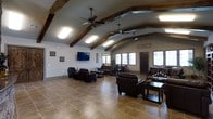 This single-floor space makes a strong architectural impression through its vaulted ceilings adorned with prominent rustic wood beams that run the length of the room, adding warmth and character to an otherwise modern interior. Dual ceiling fans are mounted along the vault, ensuring comfortable airflow throughout the generous open-plan layout. The ceiling also incorporates multiple skylight-style fixtures that supplement the natural light streaming in from a row of windows along the far wall, keeping the interior bright and welcoming throughout the day.

The flooring consists of large-format neutral stone-look tile, providing a durable and cohesive foundation across the entire space. The main area is arranged with multiple distinct seating groupings, each featuring dark leather sofas, armchairs, and low coffee tables — allowing the space to accommodate both intimate conversations and larger gatherings simultaneously. A wall-mounted flat-screen television anchors one section of the room, making it suitable for presentations or entertainment. Decorative wall art and subtle accents complement the rustic-meets-contemporary aesthetic. The overall ambiance is open, functional, and inviting, well-suited for communal or multipurpose use.