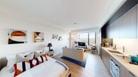 This thoughtfully designed studio unit presents a seamless open-concept layout that maximizes every square foot. Light hardwood flooring flows continuously throughout, unifying the sleeping, living, and kitchen areas into one cohesive space.

The living area is anchored by a large floor-to-ceiling window that floods the interior with natural light and frames impressive panoramic views of the surrounding urban landscape, including city buildings and distant open terrain. This corner of the unit is ideal for relaxing or entertaining, with ample room for a full seating arrangement and media setup.

The integrated kitchen runs along one full wall and features warm wood-grain cabinetry extending from floor to ceiling, a built-in oven and microwave, an undermount sink, and a clean light-toned countertop. The cabinetry design is both functional and visually striking, with dark matte hardware providing a modern contrast.

The sleeping area is positioned opposite the kitchen and is bright and airy, benefiting from the same natural light that fills the rest of the unit. A short hallway connects the main living space to the entry, where additional storage solutions are integrated into the cabinetry wall.

Clean white walls, recessed lighting, and a minimalist architectural palette give the unit a contemporary, polished ambiance throughout.