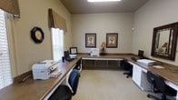 This single-story space is designed as a well-equipped business center, offering a practical and organized environment for productivity. The room features continuous built-in wooden workstations that wrap along multiple walls, providing generous desk space for multiple users simultaneously. Each station is outfitted with computers, printers, and other essential office equipment, making the space fully functional for a variety of tasks.

The neutral beige walls create a calm, professional atmosphere, complemented by light-colored carpeting that adds warmth underfoot. Vaulted ceilings contribute a sense of openness, while a window fitted with horizontal blinds and a decorative fabric valance allows natural light to filter in. Overhead fluorescent lighting ensures the space remains well-lit throughout.

Decorative touches include two framed botanical-style artworks displayed symmetrically on the main wall, a traditional analog wall clock, and a small accent lamp on the central workstation, adding a touch of warmth to the otherwise functional setting. Ergonomic rolling chairs are positioned at each workstation, prioritizing comfort during use. Overall, the space balances practicality with a welcoming, neatly appointed aesthetic.