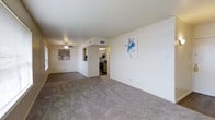 This single-floor residence offers a bright, open layout that connects the main living area to the kitchen through an arched passageway, creating a natural sense of flow throughout the space. The living area is finished with plush, neutral-toned carpeting that extends across the room, contributing to a warm and comfortable atmosphere. Smooth white walls provide a clean, versatile backdrop, accented by a pair of decorative art pieces that add subtle character to the space.

A ceiling fan with a light fixture is centrally positioned overhead, offering both ventilation and ambient lighting. A visible HVAC vent ensures year-round climate control. Natural light filters in through a window on the far wall, brightening the interior without overwhelming it.

The arched opening leads directly into the kitchen, which features dark cabinetry, a black range with overhead microwave, and tile flooring — offering a practical contrast to the soft finishes of the living area. The layout encourages easy movement between spaces, making the home well-suited for everyday living. Overall, the residence presents a clean, functional, and inviting environment throughout.