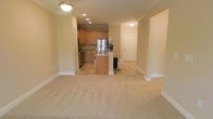 This single-floor space is designed with an open-concept layout that seamlessly connects the living area to the kitchen. The living area is finished with plush, neutral-toned carpeting that adds warmth and comfort underfoot. Crown molding runs along the upper perimeter of the space, lending a polished, refined aesthetic throughout.

The kitchen is situated toward the rear of the open layout and features honey-toned wood cabinetry, granite-style countertops, and stainless steel appliances, including a refrigerator and range. A center island with a sink provides additional workspace and serves as a natural dividing point between the kitchen and living areas. Recessed lighting in the kitchen ceiling ensures the workspace is well-lit, complemented by a pendant light fixture in the adjoining area.

The flooring transitions from tile in the kitchen and entryway to carpet in the living zone, clearly defining each functional area. A front entry door is visible in the background, positioned off a tiled foyer. The overall color palette is warm and neutral, creating a cohesive and inviting ambiance throughout the space.