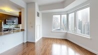 Located at 7 DeKalb in the heart of Brooklyn's Downtown district, this apartment offers a clean, contemporary aesthetic throughout its single-floor layout. Warm wood-look flooring runs continuously across the living areas and bedrooms, contrasting beautifully with crisp white walls and ceilings that maximize the sense of light and space.

The living area is generously proportioned, featuring large corner windows that frame striking views of the New York City skyline, flooding the room with natural light. Structural beam detailing on the ceilings adds an architectural dimension to the open interior. The kitchen is outfitted with stainless steel appliances, including a full-size refrigerator and range, complemented by dark cabinetry and a window overlooking the cityscape.

The bedrooms are well-sized, each featuring the same consistent hardwood-style flooring and white walls, with clean-lined doors and ample wall space. Both bathrooms are finished with large-format brown subway tile wainscoting, marble-style tile flooring, undermount sinks set into modern vanities with mirrored medicine cabinets, and chrome fixtures. One bathroom includes a walk-in glass-enclosed shower, while the other features a full soaking tub with overhead shower. Together, these spaces offer a polished, move-in ready residence with impressive urban views.