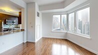Located at 7 DeKalb in the heart of Brooklyn's Downtown district, this apartment offers a clean, contemporary aesthetic throughout its single-floor layout. Warm wood-look flooring runs continuously across the living areas and bedrooms, contrasting beautifully with crisp white walls and ceilings that maximize the sense of light and space.

The living area is generously proportioned, featuring large corner windows that frame striking views of the New York City skyline, flooding the room with natural light. Structural beam detailing on the ceilings adds an architectural dimension to the open interior. The kitchen is outfitted with stainless steel appliances, including a full-size refrigerator and range, complemented by dark cabinetry and a window overlooking the cityscape.

The bedrooms are well-sized, each featuring the same consistent hardwood-style flooring and white walls, with clean-lined doors and ample wall space. Both bathrooms are finished with large-format brown subway tile wainscoting, marble-style tile flooring, undermount sinks set into modern vanities with mirrored medicine cabinets, and chrome fixtures. One bathroom includes a walk-in glass-enclosed shower, while the other features a full soaking tub with overhead shower. Together, these spaces offer a polished, move-in ready residence with impressive urban views.