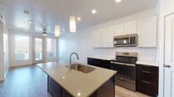 This thoughtfully designed single-floor residence offers a clean, contemporary aesthetic throughout. The heart of the home is an open-concept kitchen, showcasing a large center island with a sleek quartz countertop, an undermount sink, and elegant pendant lighting overhead. Crisp white upper cabinetry contrasts beautifully with dark lower cabinets, while a classic white subway tile backsplash ties the look together. A full suite of stainless steel appliances — including a range, over-the-range microwave, refrigerator, and dishwasher — completes the kitchen's modern appeal. Light wood-look flooring flows throughout the main living areas, enhancing the bright and airy atmosphere created by recessed lighting and natural light from generous windows. The living area features a ceiling fan and access to what appears to be a patio or balcony. The bedroom is a comfortable, carpeted retreat with a ceiling fan, ample natural light, and a neutral color palette. The bathroom is finished with wood-look flooring, a dark-cabinet vanity with a white countertop, a large mirror, and direct access to a walk-in closet with built-in shelving. A dedicated laundry space is also present, adding everyday convenience to this well-appointed home.