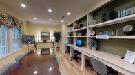Located at 1955 Poppy Ferry Rd in Biloxi, Mississippi, this well-appointed single-floor community room combines functional amenities with classic, refined décor. The space centers around a polished wood conference table surrounded by cushioned chairs, making it well-suited for meetings or collaborative gatherings. Light hardwood flooring runs throughout, adding warmth and continuity to the room.

Along one wall, a built-in shelving unit with a designated laptop area provides a practical workstation complete with desktop computers, reference books, and decorative greenery accents set against a deep sage-green backdrop. Crown molding and recessed lighting lend a polished, formal character to the interior.

Tall windows dressed with layered fabric valances and wood blinds line the exterior wall, allowing generous natural light to filter in while offering views of the surrounding outdoor greenery. The warm yellow walls create an inviting, comfortable atmosphere throughout.

A decorative console table with an ornate metal base, flanked by matching table lamps and topped with a framed landscape painting, serves as an elegant focal point in one corner. Nearby, a display board showcases property imagery, adding a community-oriented touch. French doors connect this space to an adjacent area, enhancing the overall flow and openness of the layout.