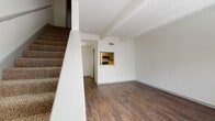 This two-story residence offers a functional and straightforward layout designed for comfortable everyday living. The main level features rich, dark hardwood-style flooring throughout the open living area, which is anchored by distinctive exposed ceiling beams that add architectural character. A carpeted staircase leads to the upper floor, while a convenient pass-through window connects the living space to the kitchen, maintaining an open feel between the two areas. Natural light fills the living room through blinds-fitted windows, creating a bright and airy atmosphere.

The galley-style kitchen is well-equipped with gray cabinetry, granite-look countertops, a double sink, and black appliances including a range and refrigerator, all set on warm wood-tone flooring. A pass-through opening to the living area enhances the sense of connectivity.

Upstairs, the carpeted bedroom provides a quiet retreat and includes a walk-in closet with built-in rod storage, along with a window that invites natural light. The bathroom is clean and practical, outfitted with a tub-shower combination, chrome fixtures, a wall-mounted gray storage cabinet, and wood-tone flooring, completing the home's cohesive design aesthetic throughout both levels.