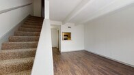 This two-story residence offers a functional and straightforward layout designed for comfortable everyday living. The main level features rich, dark hardwood-style flooring throughout the open living area, which is anchored by distinctive exposed ceiling beams that add architectural character. A carpeted staircase leads to the upper floor, while a convenient pass-through window connects the living space to the kitchen, maintaining an open feel between the two areas. Natural light fills the living room through blinds-fitted windows, creating a bright and airy atmosphere.

The galley-style kitchen is well-equipped with gray cabinetry, granite-look countertops, a double sink, and black appliances including a range and refrigerator, all set on warm wood-tone flooring. A pass-through opening to the living area enhances the sense of connectivity.

Upstairs, the carpeted bedroom provides a quiet retreat and includes a walk-in closet with built-in rod storage, along with a window that invites natural light. The bathroom is clean and practical, outfitted with a tub-shower combination, chrome fixtures, a wall-mounted gray storage cabinet, and wood-tone flooring, completing the home's cohesive design aesthetic throughout both levels.
