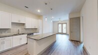 This well-appointed single-floor residence showcases a clean, modern design with thoughtful finishes throughout. The open-concept kitchen is a standout feature, outfitted with crisp white shaker cabinetry, granite countertops, a marble-style tile backsplash, and a full suite of stainless steel appliances including a refrigerator, range, microwave, and dishwasher. A generously sized kitchen island with a matching granite surface provides additional prep space and serves as a natural gathering point. Pendant lighting above the island adds a contemporary touch.

The kitchen flows seamlessly into the living area, which is bathed in natural light from a set of glass French doors that open to an outdoor balcony. A ceiling fan overhead adds comfort and airflow to the space. Durable wood-look flooring runs throughout the main living areas, lending a warm and cohesive aesthetic.

The bedroom is carpeted for comfort and features a large window that invites natural light. It connects directly to a well-designed bathroom, complete with a granite-topped vanity, a glass-enclosed shower, wood-look flooring, and direct access to a spacious walk-in closet with built-in shelving. The overall palette of neutral tones and modern hardware creates a clean, inviting ambiance throughout the residence.