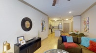 This single-floor residence offers a thoughtfully designed open-concept layout that seamlessly connects the living area and kitchen, creating a comfortable and functional living environment. The living space features light-toned flooring, crown molding, and a ceiling fan that adds both style and practicality. The area is bright and welcoming, with recessed lighting complementing the overall warmth of the space. A stylish media console and decorative wall art enhance the living area's contemporary aesthetic.

The kitchen is well-equipped with white cabinetry, stainless steel appliances including a refrigerator, stove, and microwave, and a central island with pendant lighting overhead. The island provides additional seating with bar-height stools, making it ideal for casual dining or entertaining. The clean, neutral countertops and light cabinetry give the kitchen a fresh, modern feel.

The open flow between the living and kitchen areas makes the space feel expansive and airy. Crown molding carries throughout, adding an elegant architectural detail that ties the rooms together. The entry door is visible from the main living area, reinforcing the single-floor, open design. Overall, this residence combines modern finishes with a practical layout to deliver a comfortable and inviting atmosphere.