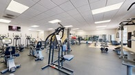 Located at 405 Greenbriar Dr in Normal, IL, this fitness facility offers a thoroughly equipped, single-floor exercise space designed to support a wide range of workout routines. The facility is organized into distinct functional zones, each catering to different training needs.

The cardio area features multiple treadmills, elliptical machines, and stationary bikes, all positioned beneath wall-mounted televisions for an engaging workout experience. Adjacent to this zone, a dedicated strength training area houses a variety of cable machines, multi-functional weight stations, and free weights including barbells and medicine balls, all neatly arranged along mirrored walls that help maximize the sense of space.

A separate functional training zone features light-colored tile flooring, a flat workout bench, and ample open floor space ideal for stretching, bodyweight exercises, or barbell work. Full-length mirrors run along the walls, enhancing visibility and form-checking during workouts.

Throughout the facility, durable rubber flooring provides safety and comfort underfoot, while a drop ceiling with recessed panel lighting ensures bright, even illumination. Natural light filters in through several windows offering views of the surrounding area. The clean, neutral color palette and well-organized layout create a focused, professional atmosphere well-suited for both individual and group fitness activities.