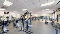 Located at 405 Greenbriar Dr in Normal, IL, this fitness facility offers a thoroughly equipped, single-floor exercise space designed to support a wide range of workout routines. The facility is organized into distinct functional zones, each catering to different training needs.

The cardio area features multiple treadmills, elliptical machines, and stationary bikes, all positioned beneath wall-mounted televisions for an engaging workout experience. Adjacent to this zone, a dedicated strength training area houses a variety of cable machines, multi-functional weight stations, and free weights including barbells and medicine balls, all neatly arranged along mirrored walls that help maximize the sense of space.

A separate functional training zone features light-colored tile flooring, a flat workout bench, and ample open floor space ideal for stretching, bodyweight exercises, or barbell work. Full-length mirrors run along the walls, enhancing visibility and form-checking during workouts.

Throughout the facility, durable rubber flooring provides safety and comfort underfoot, while a drop ceiling with recessed panel lighting ensures bright, even illumination. Natural light filters in through several windows offering views of the surrounding area. The clean, neutral color palette and well-organized layout create a focused, professional atmosphere well-suited for both individual and group fitness activities.