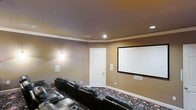 This single-level home theater is thoughtfully designed to deliver a true cinematic experience within a private residential setting. The room is anchored by a large, wall-mounted projection screen that serves as the focal point, providing an expansive viewing surface ideal for movies, sports, and entertainment. Rows of plush, black leather recliner seats are arranged in a tiered layout, ensuring unobstructed sightlines from every seat in the house. The warm, earthy tones of the walls create an intimate and immersive atmosphere, while crown molding along the ceiling adds a refined architectural touch. Recessed ceiling lighting offers subtle ambient illumination without disrupting the viewing environment, and dedicated wall-mounted speakers are strategically positioned to deliver full surround sound. The patterned area rug adds visual interest and aids in acoustic management throughout the space. A clean white six-panel door provides access to the room, maintaining a classic aesthetic that complements the warm interior palette. Every element of this home theater has been carefully considered to balance comfort, functionality, and style, making it an exceptional space for entertainment and relaxation.