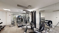 Located at 7035 Lanewood Ave in the heart of Los Angeles, CA, this fitness center offers a well-equipped workout facility across two exercise rooms on a single floor. The space is thoughtfully organized to maximize functionality within a compact footprint, providing residents or users with a comprehensive range of exercise options.

The main exercise room is lined with large floor-to-ceiling mirrors that visually expand the space and allow users to monitor their form during workouts. The room is equipped with a Star Trac treadmill, an elliptical trainer, and a multi-station cable machine for strength training. A dedicated dumbbell rack with a full range of free weights lines one mirrored wall, complemented by a flat workout bench. Recessed and flush-panel lighting provides bright, even illumination throughout. The floors are finished with a durable, textured carpet tile suitable for gym use.

The secondary exercise room extends the workout area and features additional resistance machines, including a leg extension and curl station, along with a stationary bike. A glass-paneled entry door connects the two areas, maintaining an open and accessible flow. Overhead HVAC vents ensure proper climate control, and a wall-mounted clock keeps users on track. The clean white walls and neutral tones create a focused, professional atmosphere conducive to effective training.