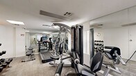Located at 7035 Lanewood Ave in the heart of Los Angeles, CA, this fitness center offers a well-equipped workout facility across two exercise rooms on a single floor. The space is thoughtfully organized to maximize functionality within a compact footprint, providing residents or users with a comprehensive range of exercise options.

The main exercise room is lined with large floor-to-ceiling mirrors that visually expand the space and allow users to monitor their form during workouts. The room is equipped with a Star Trac treadmill, an elliptical trainer, and a multi-station cable machine for strength training. A dedicated dumbbell rack with a full range of free weights lines one mirrored wall, complemented by a flat workout bench. Recessed and flush-panel lighting provides bright, even illumination throughout. The floors are finished with a durable, textured carpet tile suitable for gym use.

The secondary exercise room extends the workout area and features additional resistance machines, including a leg extension and curl station, along with a stationary bike. A glass-paneled entry door connects the two areas, maintaining an open and accessible flow. Overhead HVAC vents ensure proper climate control, and a wall-mounted clock keeps users on track. The clean white walls and neutral tones create a focused, professional atmosphere conducive to effective training.