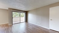 This one-bedroom, one-bathroom unit in Berkeley, CA offers a functional single-level layout encompassing a living room, bedroom, kitchen, hallway, and bathroom. The living room serves as the central gathering space, finished with wood-style laminate flooring and neutral taupe walls beneath a textured popcorn ceiling. A large sliding glass door floods the room with natural light and opens onto a private balcony overlooking mature trees, creating a connection to the outdoors. The galley-style kitchen is efficiently arranged with white cabinetry lining both walls, granite-look countertops, a tile backsplash, stainless steel gas range, and a full-size refrigerator, all illuminated by overhead lighting. A hallway connects the main living area to the bedroom and bathroom. The bedroom features plush carpeting, a window with outdoor views, a mirrored sliding closet, and a textured ceiling consistent with the rest of the unit. The bathroom includes an extended vanity with white countertops, ample cabinet storage, a large mirror, and wood-style flooring. Overall, the unit presents a cohesive, neutral palette throughout.