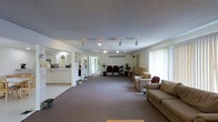 This single-floor space is designed with an open, functional layout that accommodates a variety of uses. The main area is expansive, featuring neutral-toned carpet flooring that extends throughout, contributing to a calm and approachable atmosphere. A textured ceiling with track lighting runs the length of the space, providing versatile, adjustable illumination across the room.

Along the right side, large windows with vertical blinds allow natural light to filter in, brightening the interior. The seating area along this wall includes a pair of sofas and armchairs arranged to encourage conversation and comfort.

Toward the back of the space, a row of chairs is arranged more formally, suggesting the area can serve as a meeting or waiting area. A small table with decorative floral accents adds a welcoming touch to this section.

On the left side, a built-in kitchenette is partially visible, featuring countertops, cabinetry, and basic appliances, making the space suitable for light food preparation or refreshments. A hallway extends further into the building, hinting at additional rooms or utility areas beyond the main open floor.

Overall, the space presents a clean, organized, and multi-functional environment well-suited for community, office, or service-oriented use.