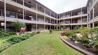 This distinctive residence is part of a two-story courtyard-style building, where units wrap around a beautifully landscaped central green space. The shared courtyard features well-maintained lawns, manicured garden beds with colorful plantings, mature trees, and ornamental decorations, creating a serene and communal outdoor atmosphere. Covered walkways with balconies line the upper level, offering residents direct views of the verdant grounds below.

Inside, the unit presents an eclectic and artistic interior defined by warm, neutral-toned walls and dark concrete flooring that adds an industrial character to the space. The dining area serves as a focal point, anchored by a striking round green marble pedestal table surrounded by woven rattan chairs, all set atop a natural woven area rug. A large-scale framed artwork with soft, abstract tones adorns the wall, contributing to the curated aesthetic. A sliding barn door with black hardware provides a stylish and functional partition between spaces, while a set of vintage-style double doors with glass panes connects to an adjoining room displaying framed artwork and accented by teal walls. Recessed lighting illuminates the interiors, and ceiling vents ensure year-round comfort. The overall ambiance blends artistic sensibility with relaxed, everyday livability.