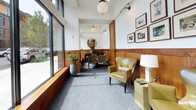 This single-floor space presents a thoughtfully designed interior that balances classic and contemporary elements. The entry corridor makes an immediate impression with its white marble-toned tile flooring adorned with a bold black Greek key mosaic border—a striking architectural detail that sets the tone for the rest of the space. Rich warm-toned wood paneling lines the walls at mid-height throughout, adding depth and a sense of traditional craftsmanship that contrasts elegantly with the white upper walls and ceiling.

The main lounge area is anchored by olive green leather wingback chairs accented with nailhead trim, positioned alongside a coordinating side table and a classic table lamp. A curated gallery of framed black-and-white photographs adorns the wall above the paneling, lending a historical and editorial quality to the space. A decorative console with a bold geometric pattern and a round mirror complete the far end of the room.

Globe pendant lights suspended from the ceiling provide warm, ambient illumination throughout. Expansive floor-to-ceiling windows along one side flood the interior with natural light and offer open views of the streetscape outside. Soft gray carpeting grounds the seating area, adding comfort underfoot and contrast to the polished tile entry. Overall, the space conveys a polished, welcoming ambiance with strong attention to material and detail.