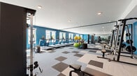 Located at 1690 North Loop in Houston, TX, this fitness center offers a spacious, single-floor layout designed to support a full range of workout routines. The interior is defined by bold blue accent walls, a crisp white ceiling with recessed lighting, and a distinctive checkerboard-patterned carpet in neutral gray and tan tones that adds visual interest while providing a durable, functional surface throughout the space. Large windows along the perimeter allow natural light to fill the room, creating an open and energizing atmosphere. Full-length mirrors are mounted along one wall, enhancing the sense of space and allowing users to monitor their form during exercise. The cardio zone features a row of treadmills, elliptical trainers, and stationary and spin bikes, while the strength training area is equipped with cable machines, a multi-functional cable tower, a rack with free weights and dumbbells, a weight bench, and plate-loaded equipment. Stability balls and additional accessories are available for functional and flexibility training. Colorful abstract artwork adorns the blue accent wall near the entrance, adding a creative touch to the practical environment. The overall layout is thoughtfully organized to provide clear circulation paths and dedicated zones for different types of training.