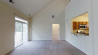 This two-story space makes an immediate impression with its dramatic vaulted ceilings in the main living area, which soar to a striking height and lend the room a sense of openness and grandeur. The living area is finished with wall-to-wall carpeting in a neutral tone, complementing the crisp, light-colored walls that enhance the brightness of the room. A large window along one wall invites natural light to fill the space, creating a warm and welcoming ambiance. A pass-through opening on one side connects the living area to the kitchen, which features warm wood-toned cabinetry and a countertop that doubles as a convenient bar or serving surface. This design choice maintains visual connectivity between the two spaces while providing functional separation. A doorway visible from the main living area leads to an adjoining room or hallway, suggesting a well-considered floor plan with thoughtful flow between spaces. Ventilation is provided by ceiling-mounted HVAC vents, ensuring comfort throughout. The overall aesthetic is clean, neutral, and versatile, offering a blank canvas well-suited to a variety of interior styles and furnishing preferences.