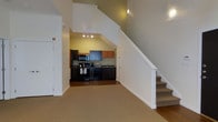 This two-story residence offers a well-organized layout across two floors, providing clear separation between living and private spaces. The main level features an open living area with neutral carpet flooring that transitions into a kitchen finished with hardwood-style flooring. The kitchen is equipped with dark cabinetry contrasted by warm wood-toned upper cabinets, black appliances including a range and built-in microwave, and a sink set beneath task lighting. A dedicated track lighting system illuminates the kitchen area effectively. The staircase, finished in crisp white with a clean balustrade, rises prominently alongside the kitchen and draws the eye upward to a vaulted ceiling, adding a strong sense of vertical space and openness to the main level. Accent lighting is recessed into the upper wall, enhancing the airy feel. The upper floor houses the two bedrooms and provides a private retreat separated from the main living areas. With two full bathrooms spread across both levels, the layout accommodates ease of use for residents and guests alike. The overall design balances functionality with a clean, contemporary aesthetic.