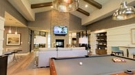 This single-floor space is designed to impress, combining upscale architectural details with a warm, inviting ambiance. The centerpiece of the main lounge area is a dramatic floor-to-ceiling stacked stone fireplace, topped with a wall-mounted flat-screen television, creating a natural focal point for the room. Soaring vaulted ceilings with exposed dark wood beams lend a sense of grandeur and open airiness, while a striking hammered metallic pendant light adds an elegant, contemporary touch overhead. The lounge is furnished with plush, neutral-toned sofas arranged to encourage conversation and relaxation. A full-size billiards table anchors the foreground of the space, offering a recreational element that complements the social atmosphere. To one side, built-in wooden shelving provides both decorative display space and functional storage, adding a refined, library-like quality to the area. Large glass partitions and windows allow natural light to flow freely throughout, connecting the interior to adjacent spaces and maintaining a bright, open feel. The overall design palette — featuring stone, wood, and soft neutrals — strikes a balance between sophistication and comfort, making this a versatile space well-suited for both leisure and social gatherings.