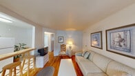 This single-floor residence offers a warm and inviting layout with a cohesive, cottage-inspired aesthetic throughout. Gleaming hardwood floors run through the main living areas, anchoring the space with natural warmth and continuity.

The living room serves as the heart of the home, featuring a plush sofa accented with decorative pillows, a clean white coffee table, and framed wall art in soft blue-grey tones. A braided area rug adds a charming, casual touch, while a side chair and table lamp create a cozy reading nook. An open sightline connects the living area to the rest of the unit, enhancing the sense of openness.

The kitchen, visible from the dollhouse view, includes a classic stovetop range and counter space suitable for everyday meal preparation. A small dining area with bar-style seating is conveniently positioned nearby.

The bedroom is tastefully appointed with light wood furniture, including a full-sized bed with a floral comforter in soft teal tones, a matching nightstand, dresser, and wardrobe. Two windows with valances allow natural light to filter in, and a wall-mounted radiator provides efficient heating. Nautical-themed framed artwork adds a subtle coastal character to the room.

The bathroom is bright and clean, featuring a tiled tub-shower combination with a floral shower curtain and a window overlooking outdoor greenery, bringing in natural light and a refreshing ambiance.