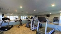 This single-floor fitness center is thoughtfully designed to support a comprehensive workout experience. The space is outfitted with a variety of cardio equipment, including elliptical trainers, treadmills, and a stationary bike, providing ample options for cardiovascular exercise. Strength training equipment, such as a weight bench and additional resistance machines, rounds out the fitness offerings. Large floor-to-ceiling mirrors line the walls, visually expanding the space and allowing users to monitor their form during exercise. The textured ceiling features recessed lighting, ensuring the room is well-lit throughout. Natural light filters in through a row of windows along one wall, adding brightness and an airy feel to the environment. Durable, low-pile carpet covers the floor, offering both comfort and stability underfoot. Wall-mounted screens are also present, providing entertainment options during workouts. The overall layout is open and organized, with equipment arranged to maximize usable floor space and allow for easy movement between stations.