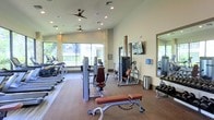 This fitness center offers a thoughtfully designed, single-floor workout facility built to accommodate a wide range of exercise needs. The space features an open layout with high ceilings and recessed lighting complemented by ceiling fans, ensuring comfortable airflow throughout. Large floor-to-ceiling windows along one wall flood the interior with abundant natural light while providing views of the surrounding outdoor area, creating an energizing atmosphere for exercise.

The cardio zone lines the windowed wall and includes a row of treadmills and elliptical machines, taking advantage of the natural light and views. The strength training area is centrally positioned and features a variety of cable and resistance machines, along with a dedicated free weights section complete with a dumbbell rack and wall-mounted mirrors for form monitoring. A weight bench is also available for versatile strength exercises.

The flooring transitions between hard surface and padded carpet zones, delineating different workout areas while providing appropriate support underfoot. A wall-mounted flat-screen television adds entertainment options for users. Decorative stone column accents and warm neutral wall tones contribute to a polished, welcoming aesthetic, making this fitness center both functional and visually appealing.