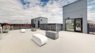 Located in the heart of Minneapolis along SE Oak Street, this rooftop terrace offers an expansive outdoor amenity space designed for both relaxation and social gathering. The deck is finished with light-toned composite decking that complements the clean, modern aesthetic of the surrounding architecture.

The terrace is thoughtfully furnished with contemporary white lounge seating, including sculptural bench-style pieces and cushioned chairs, arranged to encourage conversation and comfort. A substantial concrete fire pit serves as a central focal point, providing warmth and ambiance for evening gatherings. Additional high-top tables and chairs offer a more casual dining or socializing option.

A dedicated gas grill station is positioned along the perimeter, making the space well-suited for outdoor cooking and entertaining. The entire rooftop is enclosed by sturdy black iron fencing, ensuring both safety and unobstructed sightlines to the surrounding Minneapolis skyline and cityscape.

Transparent glass-paneled storage units are integrated into the rooftop layout, maintaining an open, organized feel. Access to the terrace is provided through a secure door from the building's interior. The open-sky exposure offers abundant natural light throughout the day, while the elevated vantage point delivers panoramic urban views that distinguish this rooftop as a standout communal amenity.