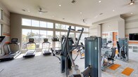 This fitness center is designed to provide a bright, motivating environment for a comprehensive workout experience. The space is characterized by soaring ceilings and expansive floor-to-ceiling windows that line multiple walls, flooding the interior with abundant natural light and offering views of the surrounding outdoor pool area and landscaped grounds. Ceiling fans are installed throughout to maintain comfortable airflow.

The cardio zone is well-stocked with a row of Matrix and other brand treadmills, elliptical trainers, and stair climbers positioned directly in front of the large windows, allowing users to exercise with an outdoor view. The strength training area features a broad selection of cable machines, weight machines, and resistance equipment arranged across a generously sized open floor plan.

The flooring consists of a distinctive patterned carpet in neutral gray tones with bold orange striped accents, complementing the room's color scheme of warm gray walls with orange horizontal banding. Recessed lighting supplements natural light throughout.

A dedicated amenity alcove near the entrance provides added convenience, featuring a compact refrigerator, folded towel service, and a pendant-lit nook framed in warm wood tones. A wall-mounted television is also present in the strength training area. Large glass entry doors complete the open, accessible design.