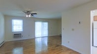 This single-floor space offers a clean, open layout highlighted by warm hardwood flooring that spans the main living area, reflecting natural light throughout the room. The space is designed with simplicity and functionality in mind, featuring neutral-toned walls that create a bright, airy atmosphere. A ceiling fan with an integrated light fixture is centrally positioned, providing both air circulation and ambient lighting. Large sliding glass doors dressed with vertical blinds serve as a focal point along one wall, allowing generous amounts of natural light to filter in while also providing access to an outdoor area. A side window further enhances the brightness of the room. Baseboard heating units are visible along the walls, indicating a practical heating solution for year-round comfort. White baseboards and trim provide clean contrast against the hardwood floors, contributing to the overall tidy aesthetic. The open floor plan offers flexible arrangement possibilities, making the space adaptable for various uses. The overall ambiance is light and neutral, providing a straightforward, move-in-ready environment with classic residential finishes.